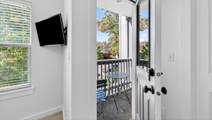 Una habitación interior con paredes blancas, un televisor montado en la pared, una puerta que da a un balcón exterior con mesa y sillas, y vistas a un área residencial con árboles y casas.