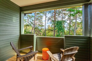 Een houten terras met twee Adirondack-stoelen en een kleine oranje kruk, met uitzicht op een bebost gebied door een raam.
