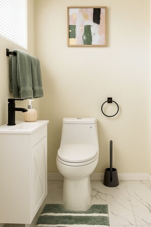Ein Badezimmer mit Toilette, Waschbecken mit Seifenspender, Handtuchhalter mit Handtüchern, Toilettenbürste und einem Bild an der Wand.