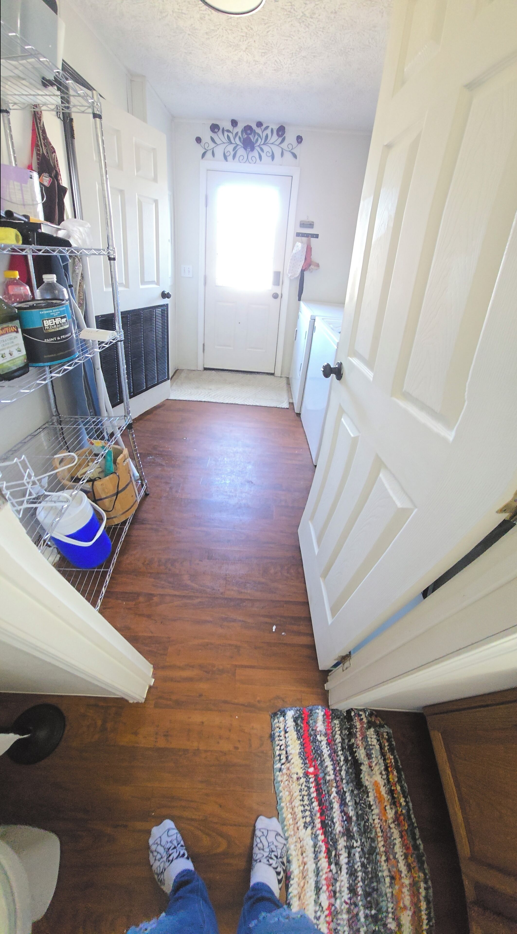 Un couloir doté d’un plancher en bois, d’un présentoir en métal contenant des produits de nettoyage, une porte agrémentée d’un élément décoratif et un tapis à l’entrée.