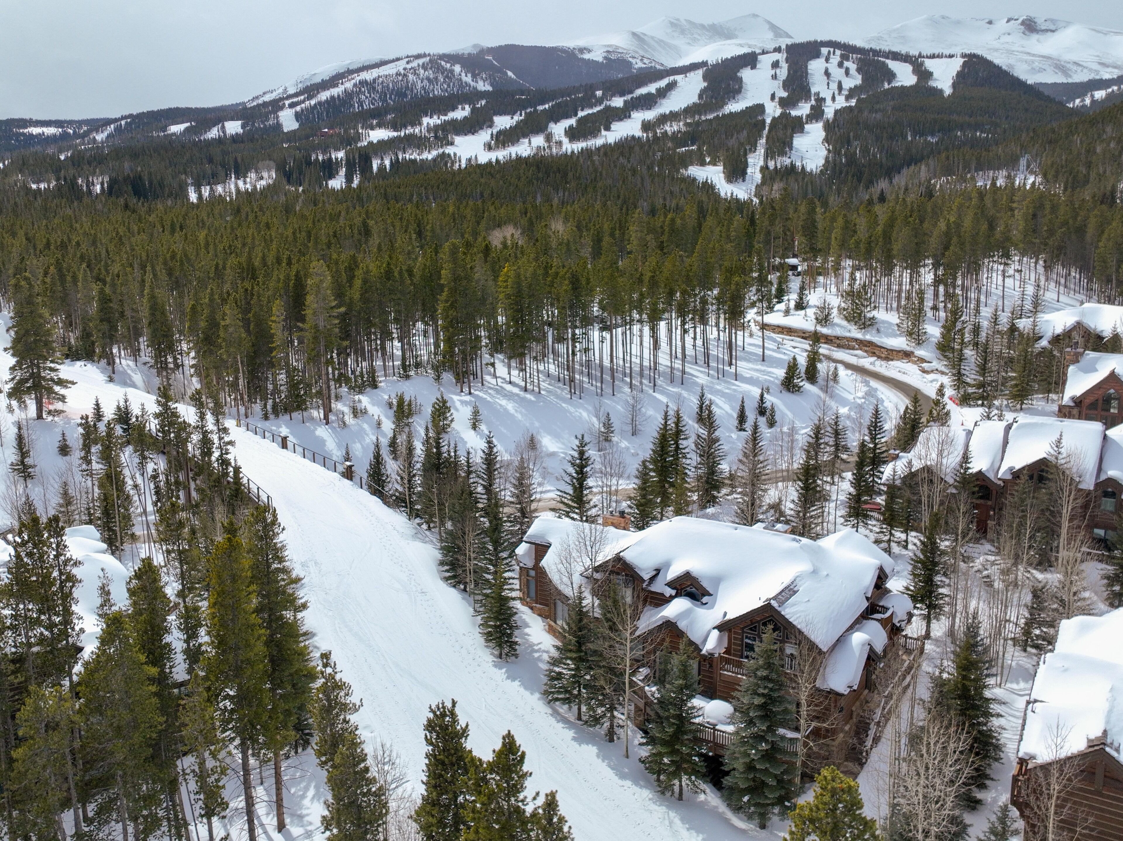 A snow-covered mountain landscape with a lodge featuring multiple wooden cabins, surrounded by dense evergreen trees.