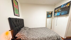 Een keurig opgemaakt bed met een grijze deken, een zwart hoofdbord en een klein nachtkastje met een lamp.