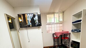 Een kleine kamer met een bed, een aan de muur bevestigde televisie, een kleintje tafel met geruit tafelkleed en een boekenplank.