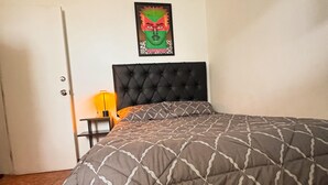 Een slaapkamer met een bed, hoofdeinde, een poster aan de muur, een deur en een klein tafeltje met een lamp.