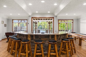 Un espace bar doté d’un comptoir en pierre, de tabourets en bois et de grandes fenêtres offrant une vue sur la verdure.