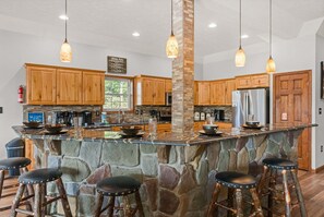 Une cuisine avec des meubles en bois, un îlot central en pierre avec des tabourets, des luminaires suspendus et un extincteur.