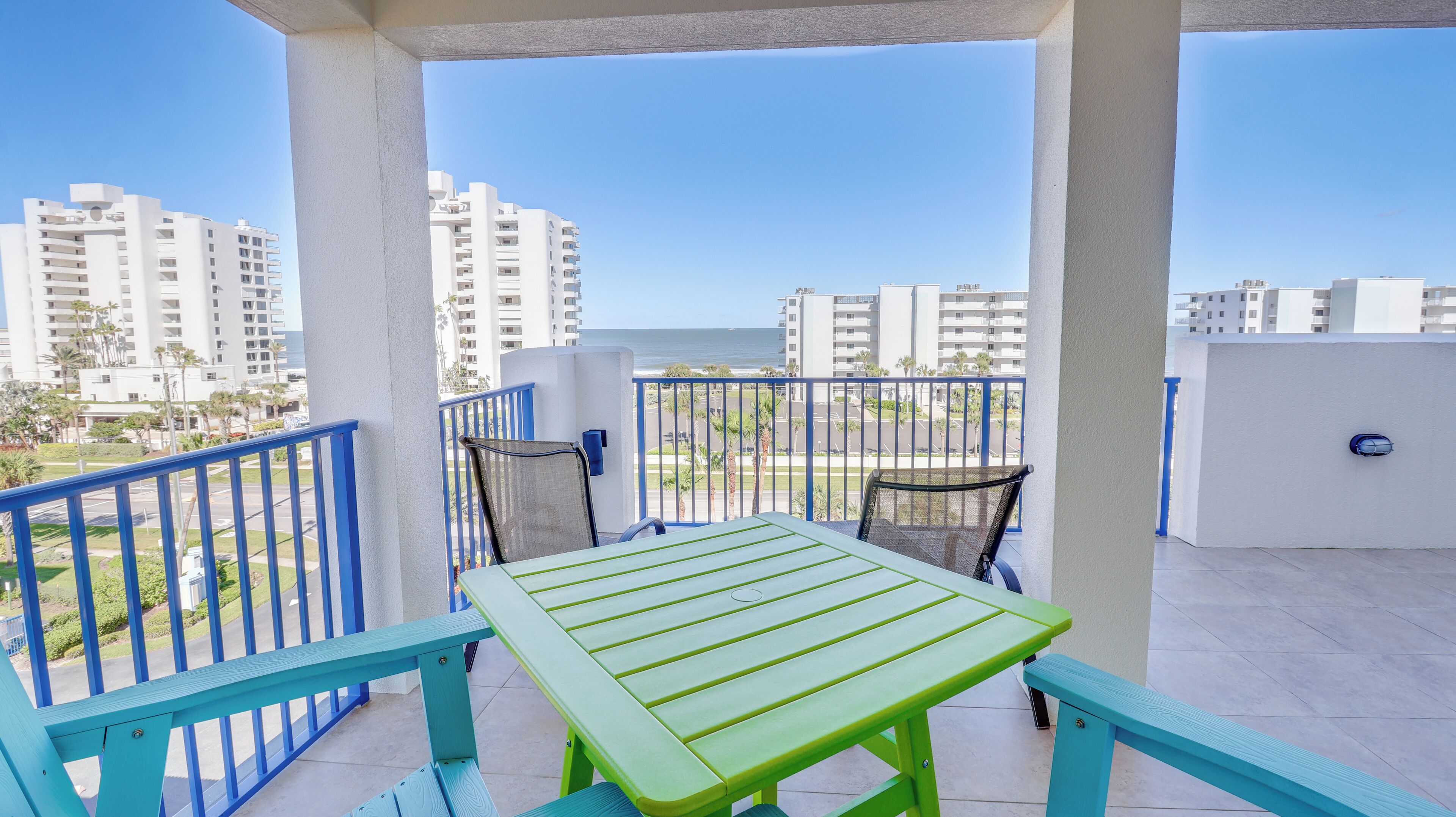 Un balcon avec une table verte et des chaises bleues, donnant sur un paysage urbain avec de hauts immeubles et un ciel dégagé.