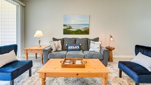 Een woonkamer met een grijze bank, twee blauwe fauteuils, een houten salontafel, en een schilderij van een strand.