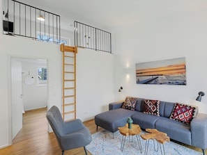Una sala de estar moderna con un sofá gris, mesitas de madera, una escalera y un cuadro de una puesta de sol.