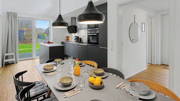 A modern kitchen with black pendant lights, a dining table set for four, and a view of the outdoors through a glass door.