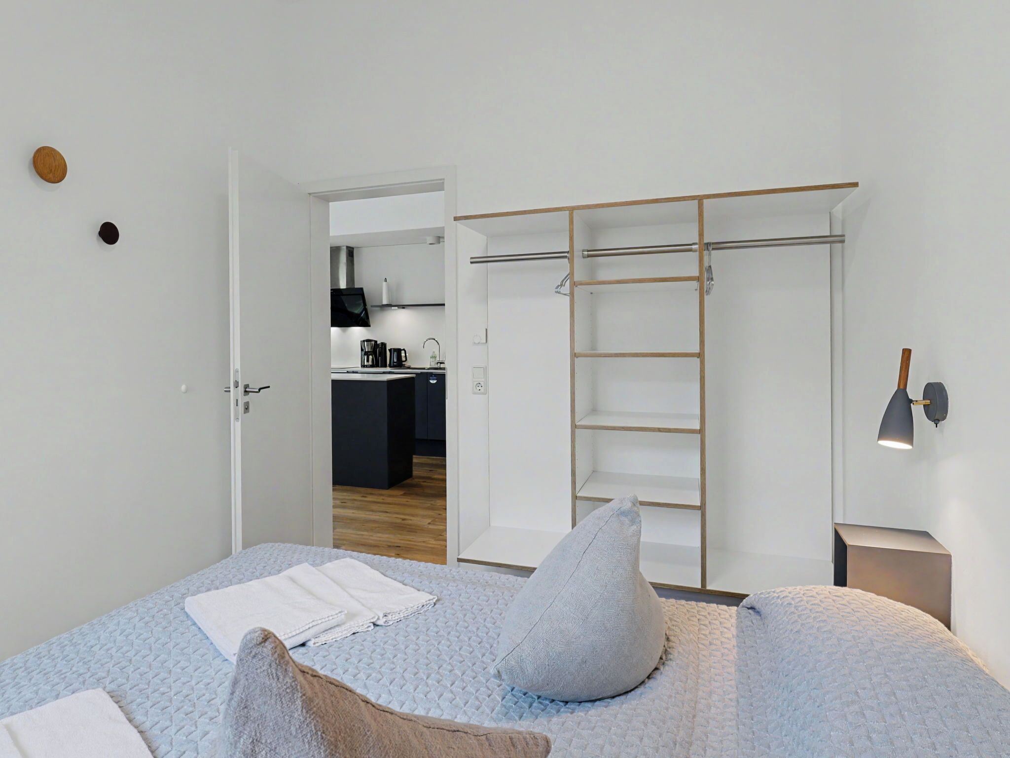 Een slaapkamer met een bed, kussens, een nachtkastje, een kledingkast en uitzicht op de keuken.