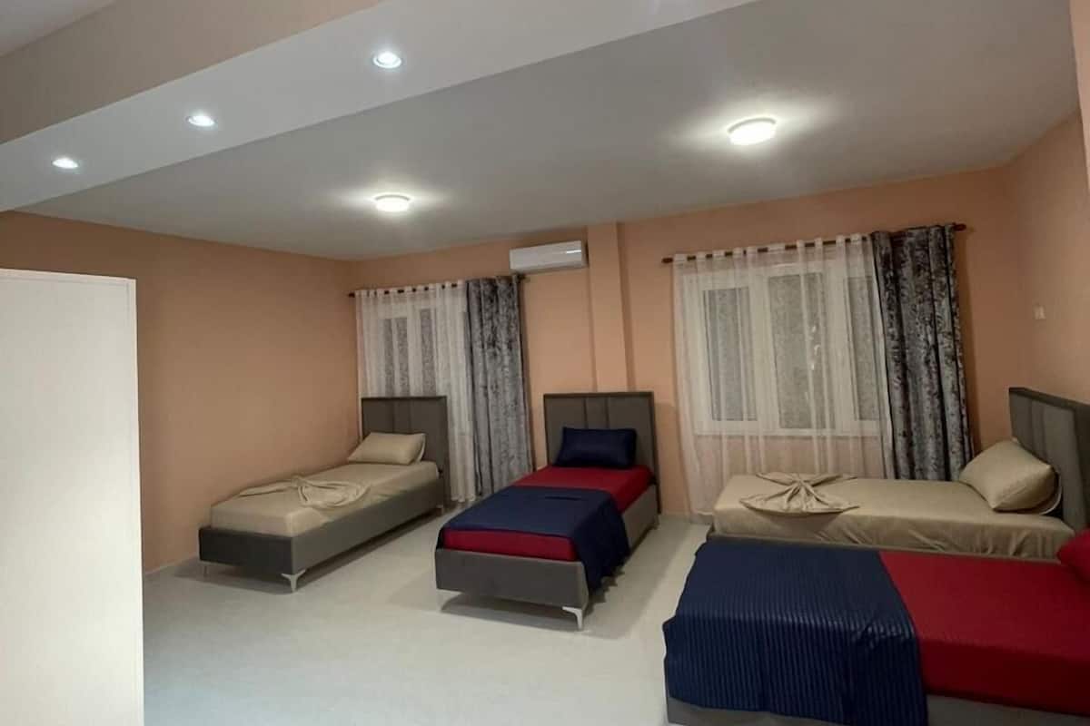 Habitación de hotel con tres camas dispuestas en fila, un techo con iluminación empotrada y una ventana con cortinas translúcidas.