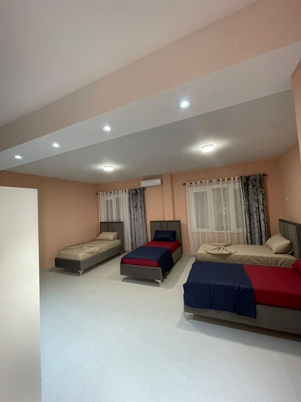 Habitación de hotel con tres camas dispuestas en fila, un techo con iluminación empotrada y una ventana con cortinas translúcidas.