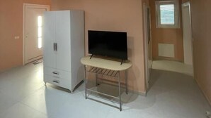 A spacious room with a white wardrobe, a TV on a stand, and a window with blinds.