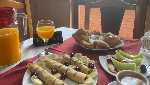 A breakfast table with a red tablecloth, a glass of orange juice, a plate of rolled crepes with bananas and cherries, a plate of croissants, a cup of coffee, and a view of greenery outside.