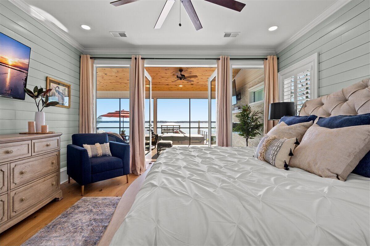Une chambre à coucher avec un grand lit, un fauteuil bleu, une commode en bois et une vue sur l’extérieur par une grande fenêtre.