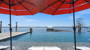 Vista a la piscina con cielo azul despejado, un muelle que se adentra en el agua y sillones de descanso bajo sombrillas rojas.