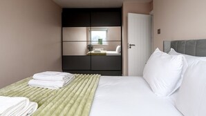 A neatly made bed with white linens and a green throw blanket, a grey upholstered headboard, and a large wardrobe with a mirror. - GuestReady - Charming 3-Bedroom London Retreat (London)