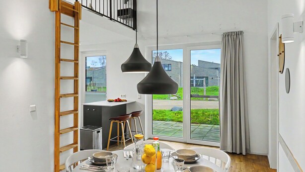 Uma sala de jantar com uma mesa redonda posta para quatro pessoas, uma cozinha ao fundo e vista para o exterior.