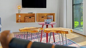 Una sala de estar con un televisor montado en la pared, una pequeña mesa con sillas y un ventanal grande.