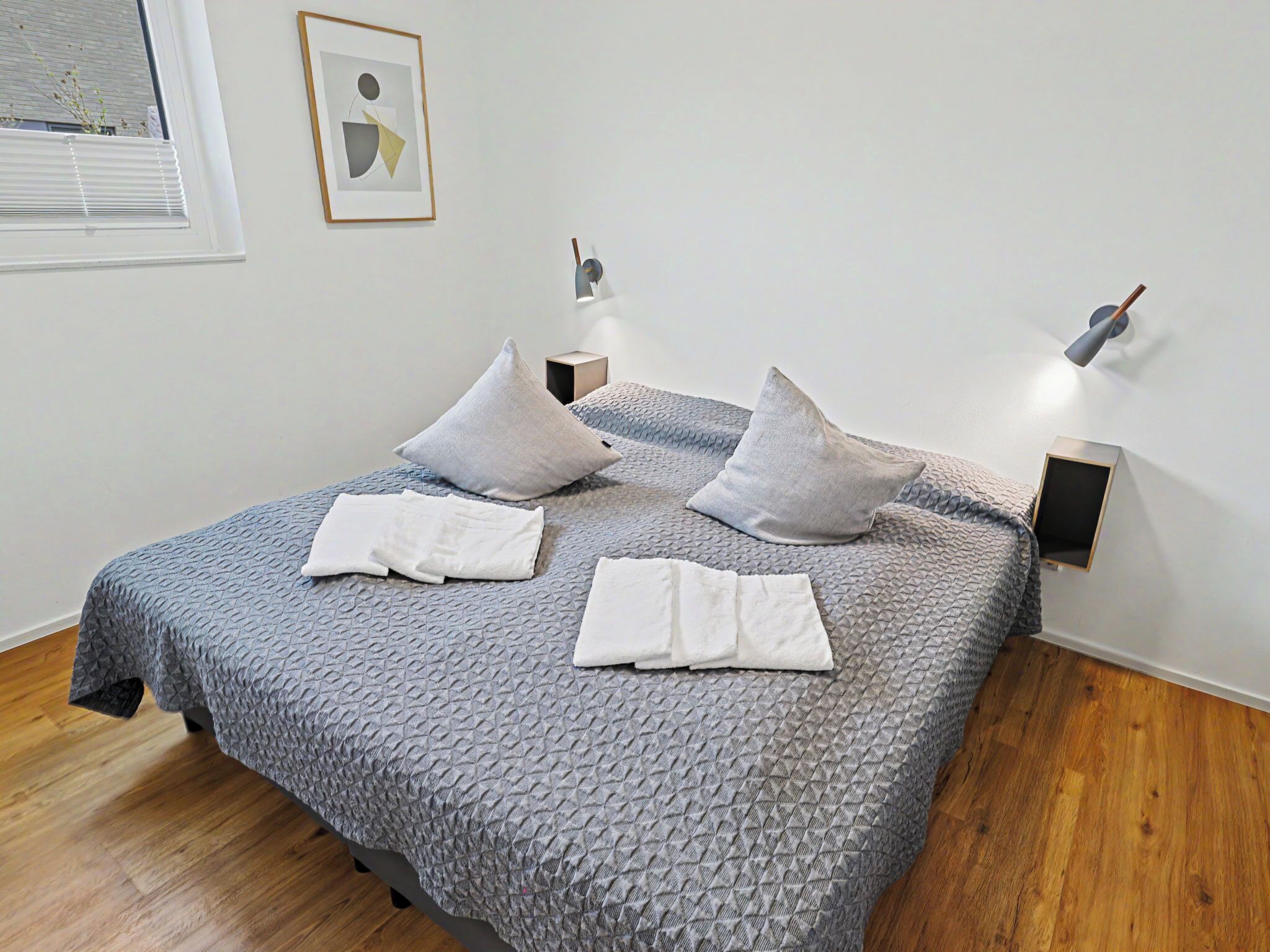 A neatly made bed with a textured blanket, two pillows, and two folded towels. There are two wall-mounted nightstands with lamps, and a framed picture on the wall.