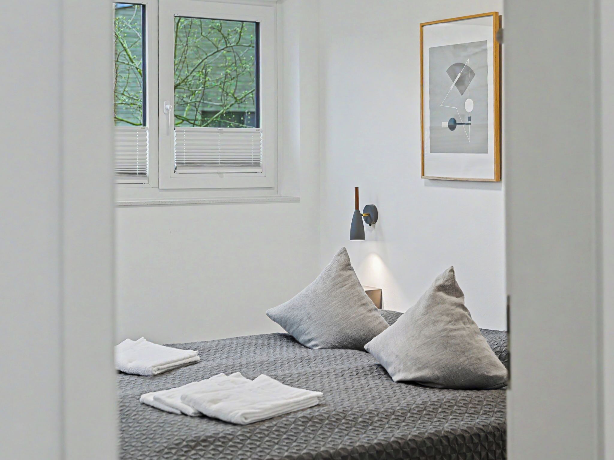 Une chambre à coucher avec un lit, des oreillers, une fenêtre avec des stores, une lampe fixée au mur et un tableau encadré.