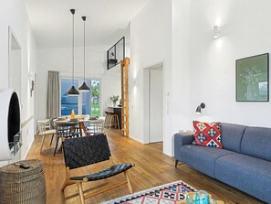 Ein modernes Wohnzimmer mit einem blauen Sofa, einem Essbereich mit Tisch und Stühlen, einem schwarzen Ledersessel und Blick ins Freie.