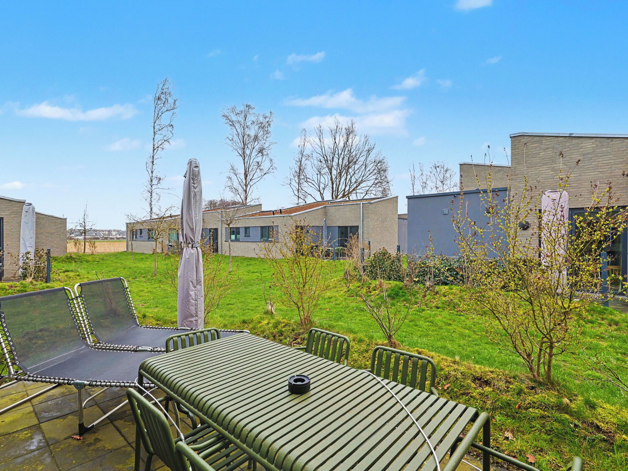 A modern outdoor seating area with metal chairs and a table, surrounded by a grassy lawn and buildings in the background.