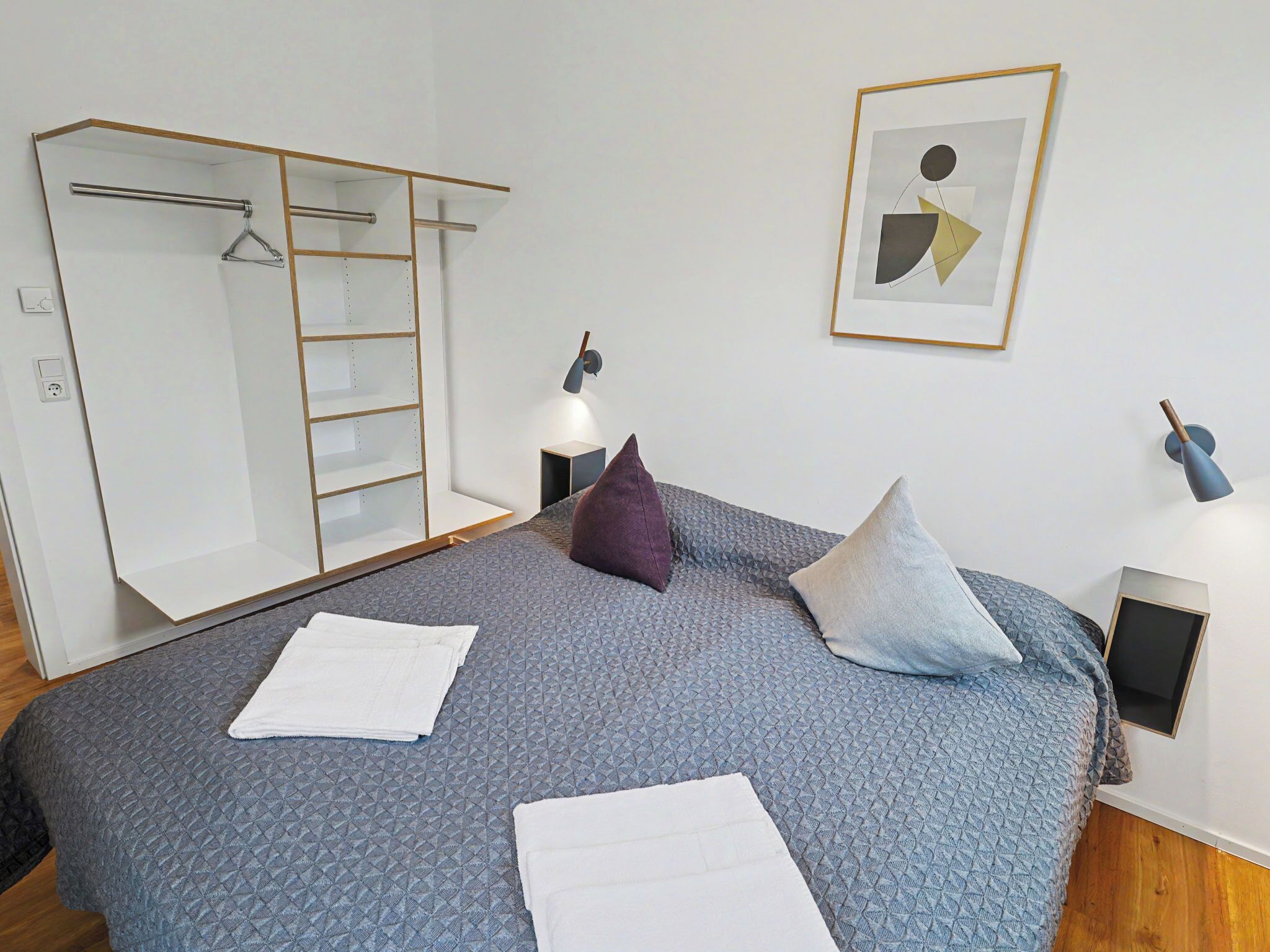 Ein Schlafzimmer mit einem Bett, einem Kleiderschrank, einer Garderobe, einem gerahmten Bild und einem Nachttisch mit einer Lampe.