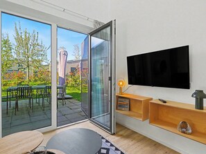 Een moderne woonkamer met een televisie, een glazen deur naar een terras met tuinmeubilair, en een klein schap met decoratieve objecten.