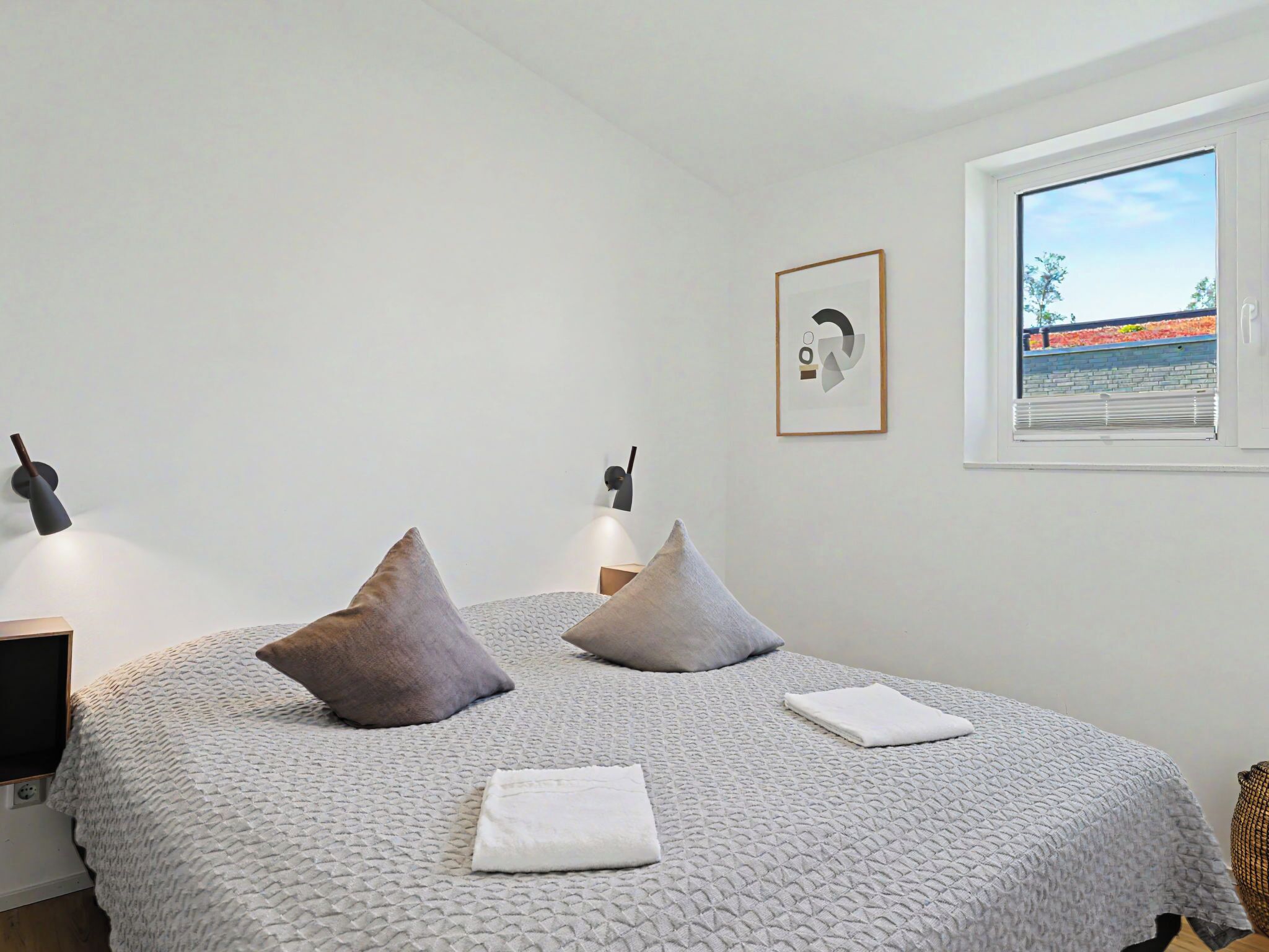 Een slaapkamer met een bed, kussens, een nachtkastje, een lamp, een foto aan de muur en een raam met uitzicht.