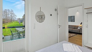 Una camera da letto con un'ampia finestra, un letto con copria letto, un comodino e vista su un giardino e edifici vicini.