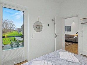 Ein Schlafzimmer mit großem Fenster, einem Bett mit Bettdecke, einem Nachttisch und Blick auf einen Garten sowie benachbarte Gebäude.