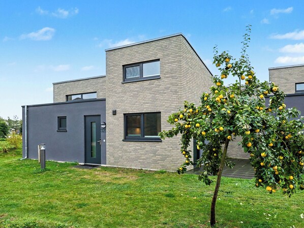 Ein modernes, zweistöckiges Gebäude mit flachem Dach und großen Fenstern. Im Vordergrund steht ein kleiner Baum mit gelben Früchten.