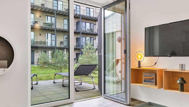 A modern apartment with a sliding glass door leading to a balcony, a flat-screen TV, a small table, and a shelf with a book and decorative items. - Goldfisch 191 (Seenähe) by Interhome (Lembruch)