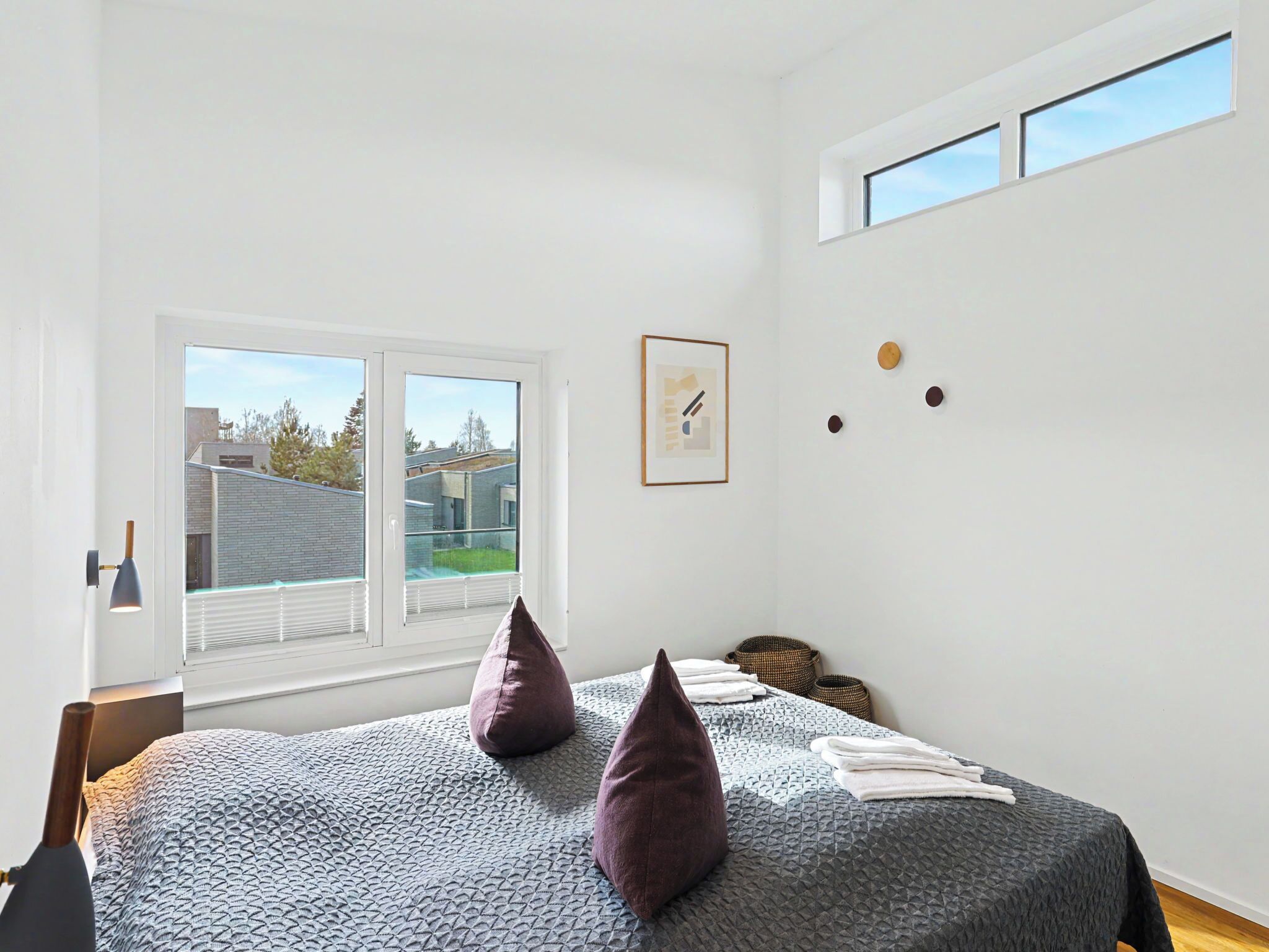 A bedroom with a large window, a bed with a textured blanket, two pillows, a bedside lamp, and a framed picture on the wall.