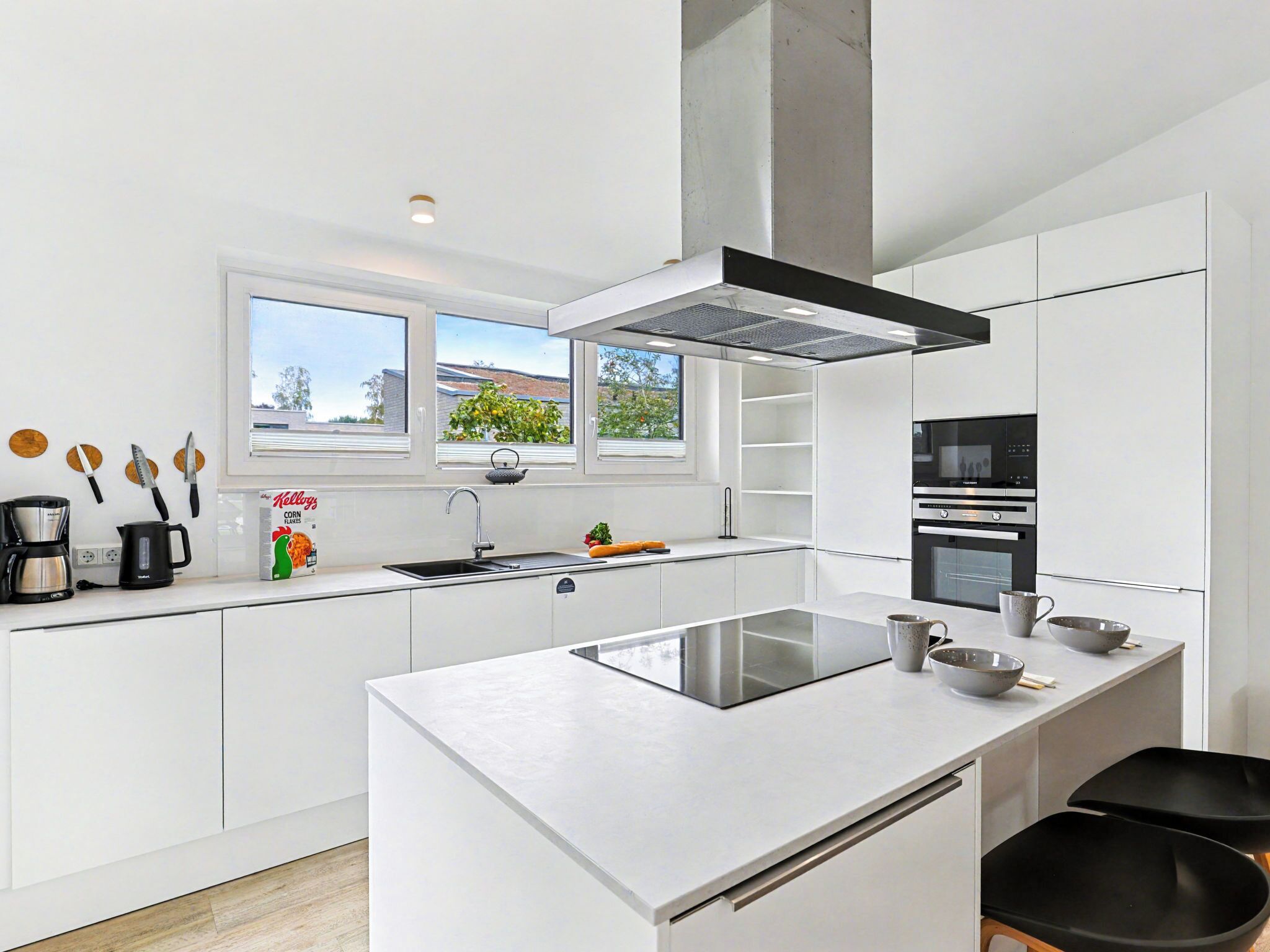 A modern kitchen with white cabinetry, a large island, a built-in oven, and a window overlooking a garden.