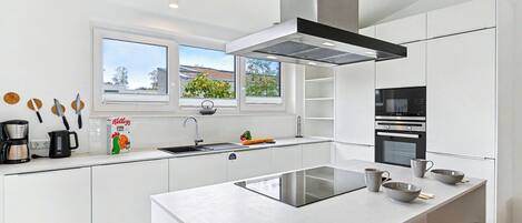 A modern kitchen with white cabinetry, a large island, a built-in oven, and a window overlooking a garden.