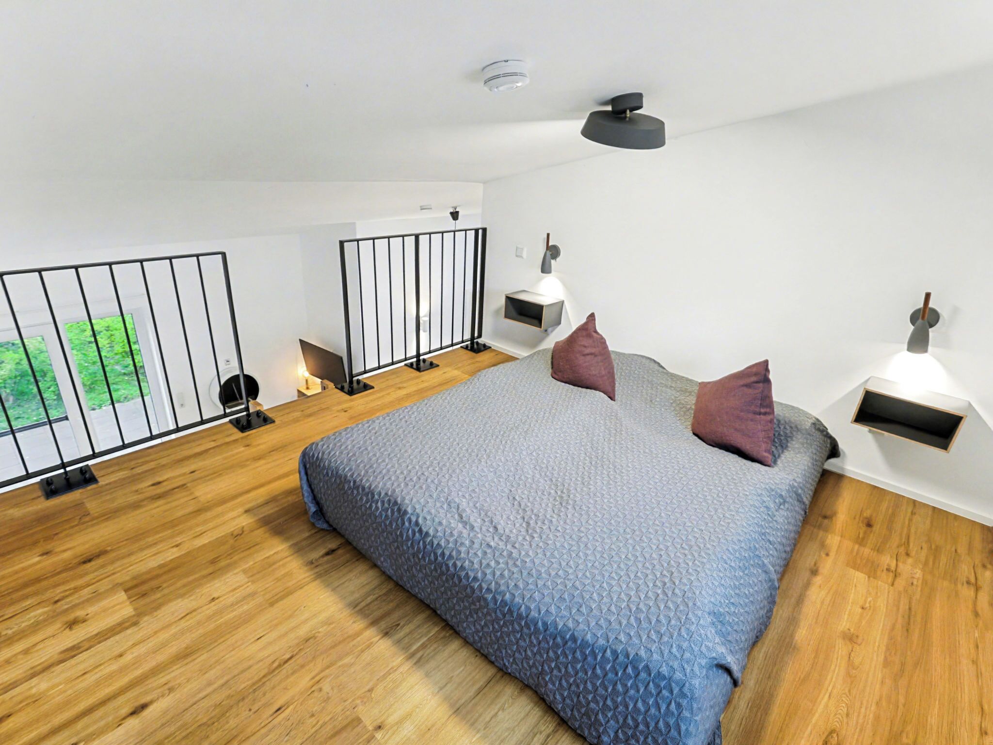 Ein modernes Schlafzimmer mit einem großen Bett, Holzboden, einem Balkon mit Metallgeländer und einem kleinen Nachttisch mit Lampe.