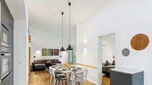Eine moderne Küche mit grauen Schränken, ein Essbereich mit einem Holztisch und -stühlen sowie ein Wohnbereich mit einer Couch und Kissen.