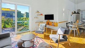 Un salon moderne doté d’un téléviseur à écran plat, d’un coin repas et d’un balcon offrant une vue sur les bâtiments et la verdure.