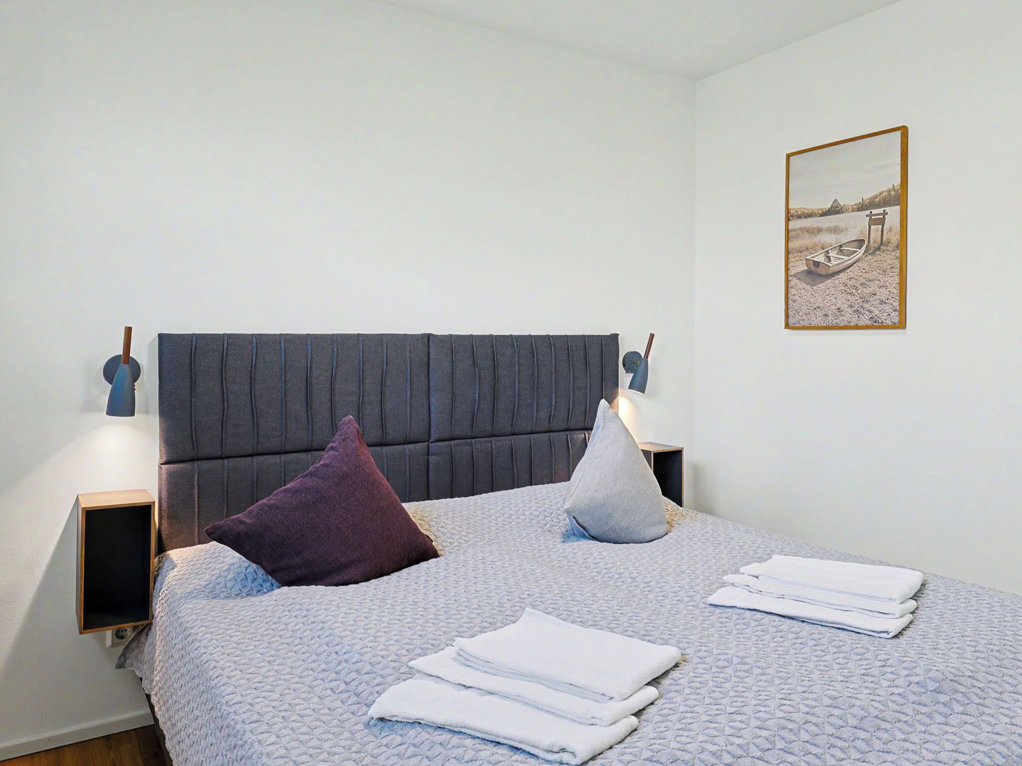 En snyggt bäddad säng med en grå sänggavel, två sänglampor, en hopvikt vit handduk och en inramad tavla på väggen.