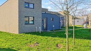 Un bâtiment moderne avec une façade en briques, un toit plat et un petit jardin devant, agrémenté d’un arbre.