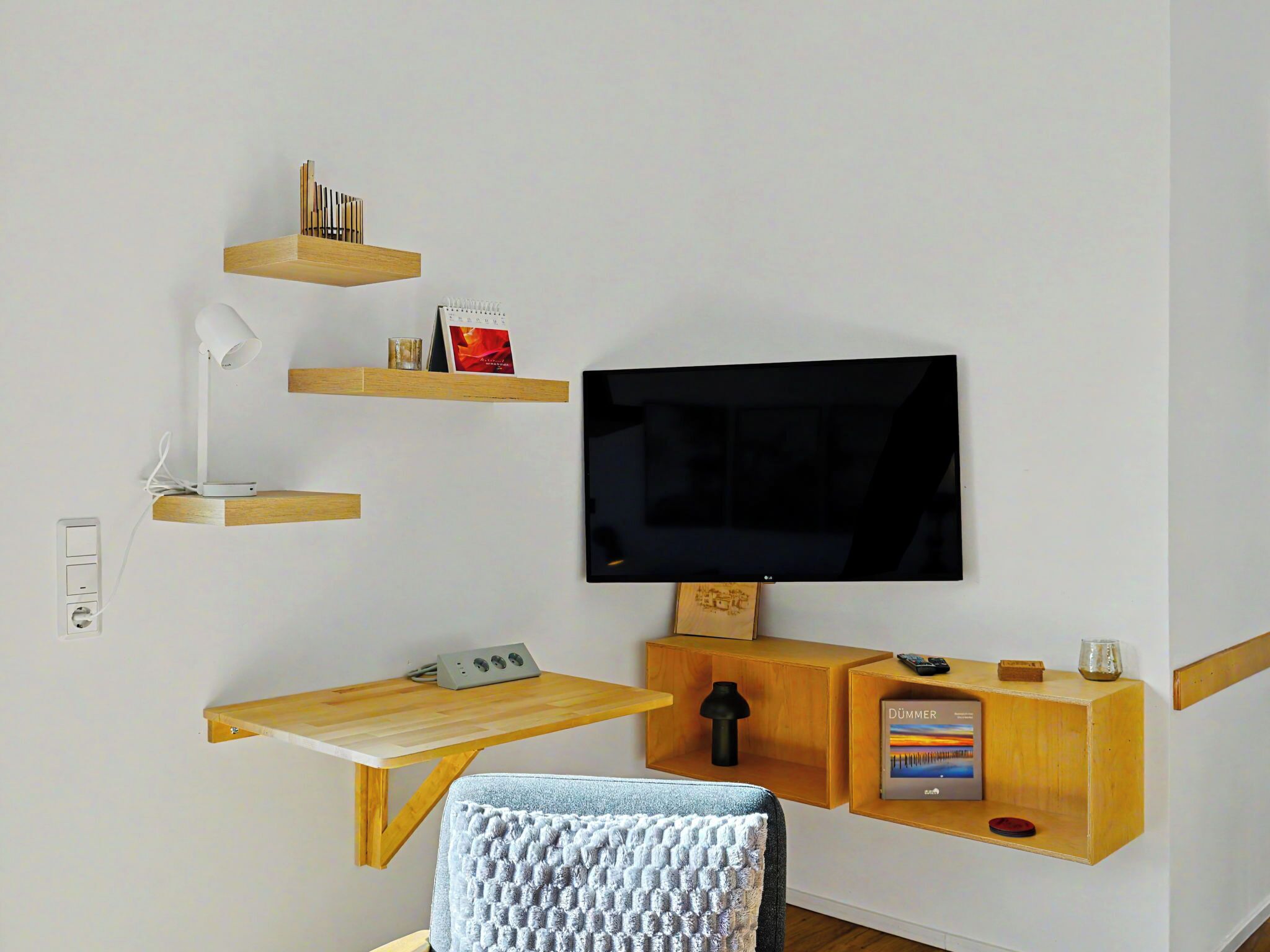 A modern living space with a flat-screen TV, wooden shelves, a desk with a lamp, and a cozy chair.