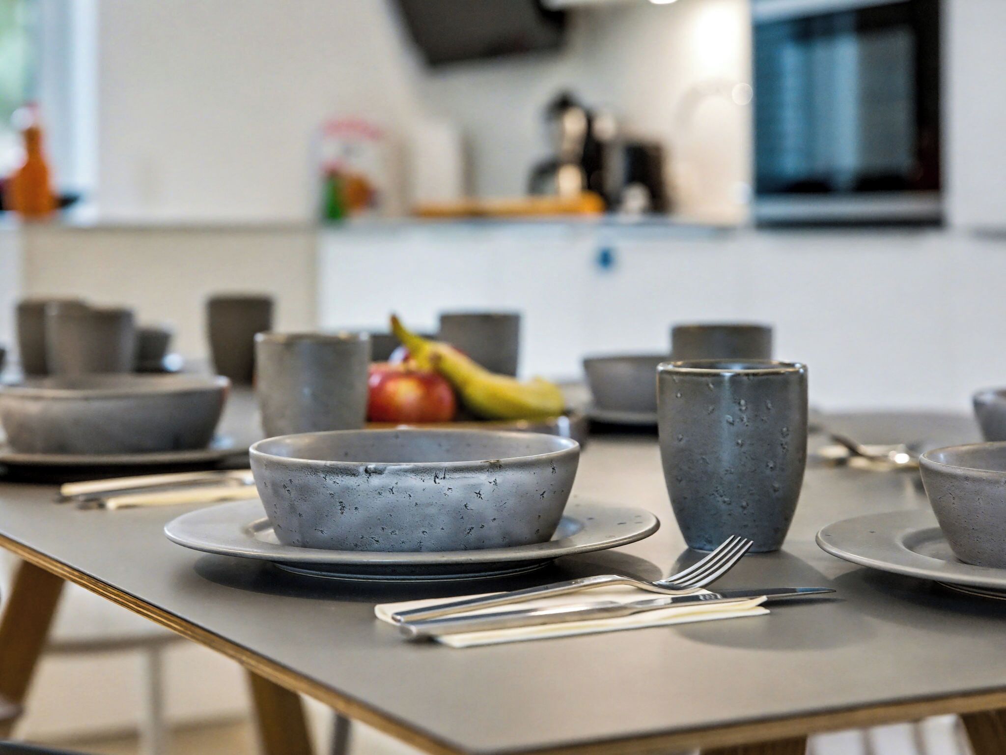 A modern kitchen with grey ceramic bowls and plates on a table, utensils, and a fruit bowl in the background.