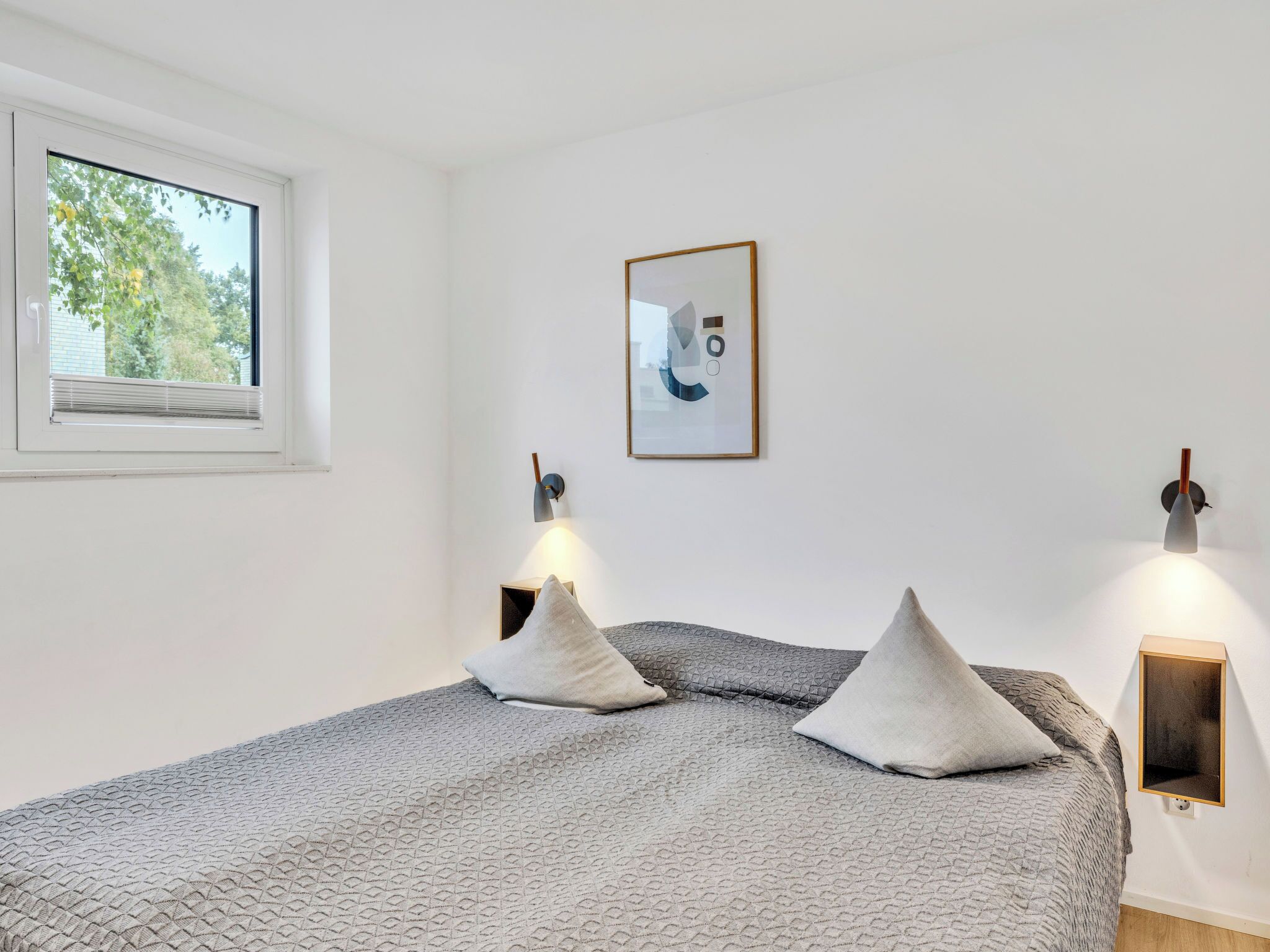 Um quarto com uma cama, duas luzes na parede, uma janela e um quadro pendurado na parede.