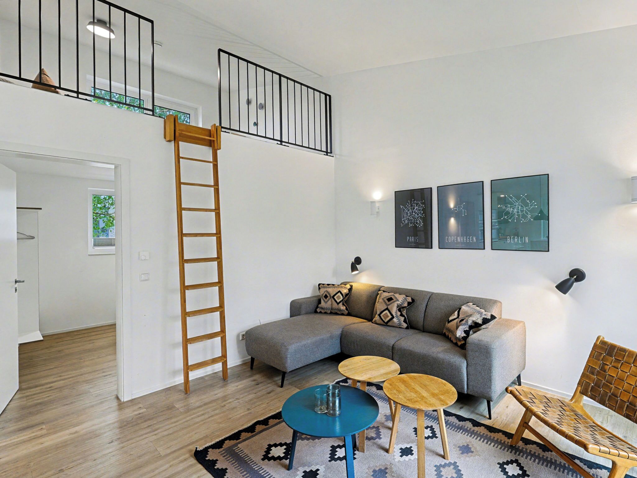 A modern living room with a grey sectional sofa, two round wooden tables, a blue coffee table, a wooden ladder, and a rattan chair.