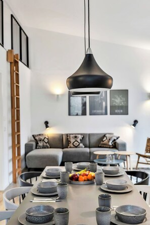 A modern dining area with a long table set for a meal, a black pendant light, and a grey sofa with cushions in the background.