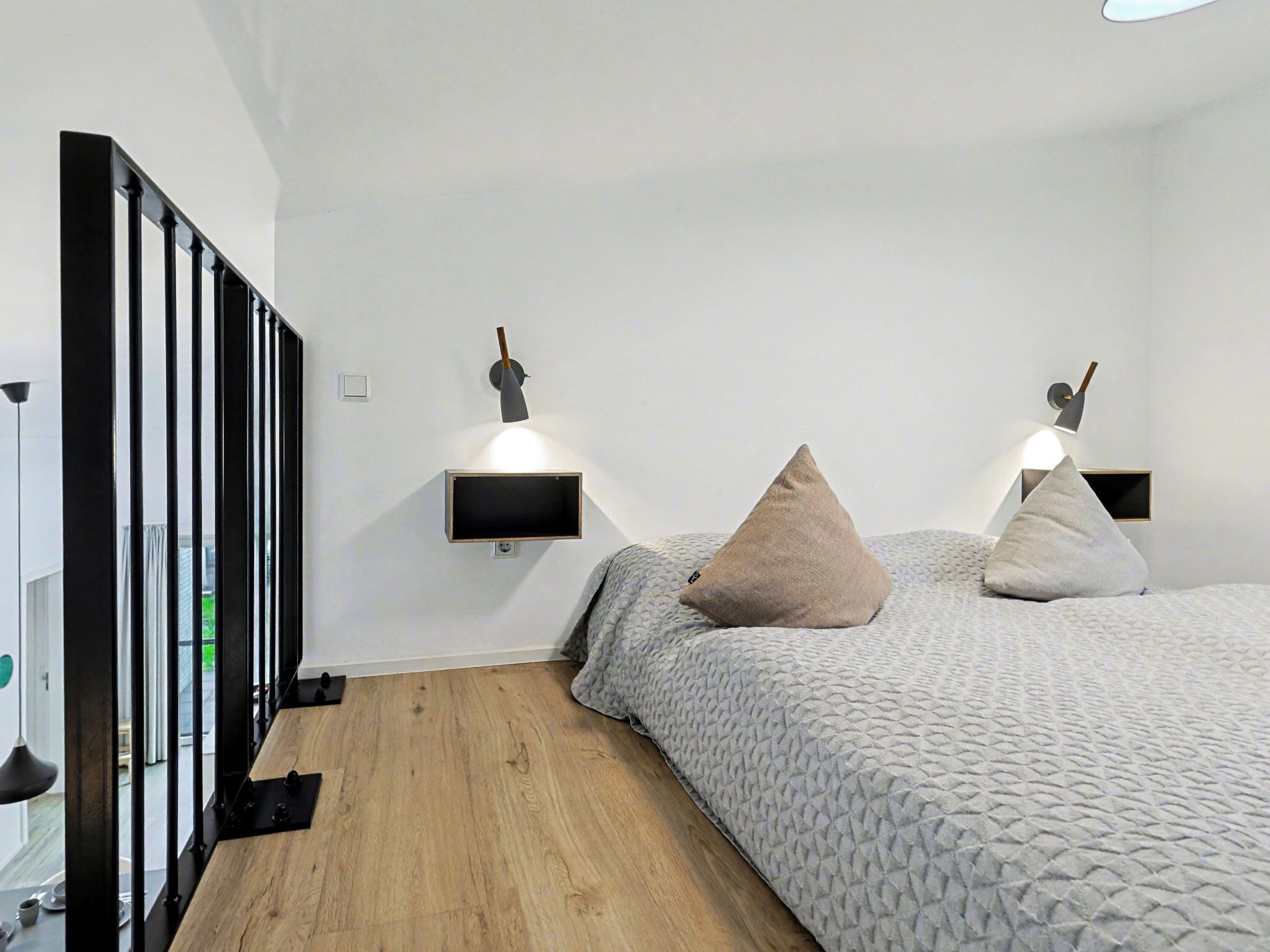 Une chambre moderne avec un plancher en bois, un lit recouvert d’une couverture à motifs, deux lampes fixées au mur et une balustrade noire.