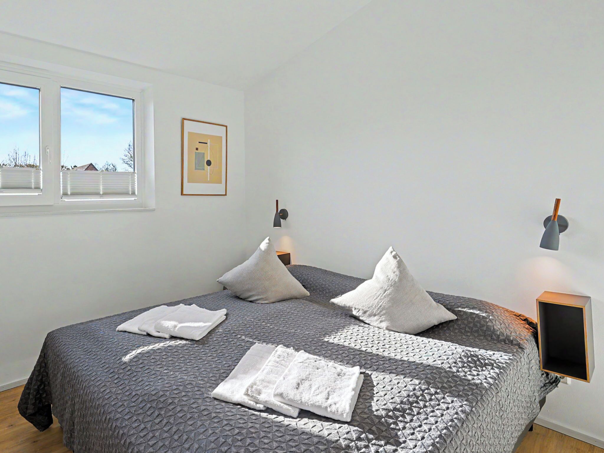 A neatly made bed with pillows and a folded towel, a wall-mounted lamp, and a framed picture on the wall.